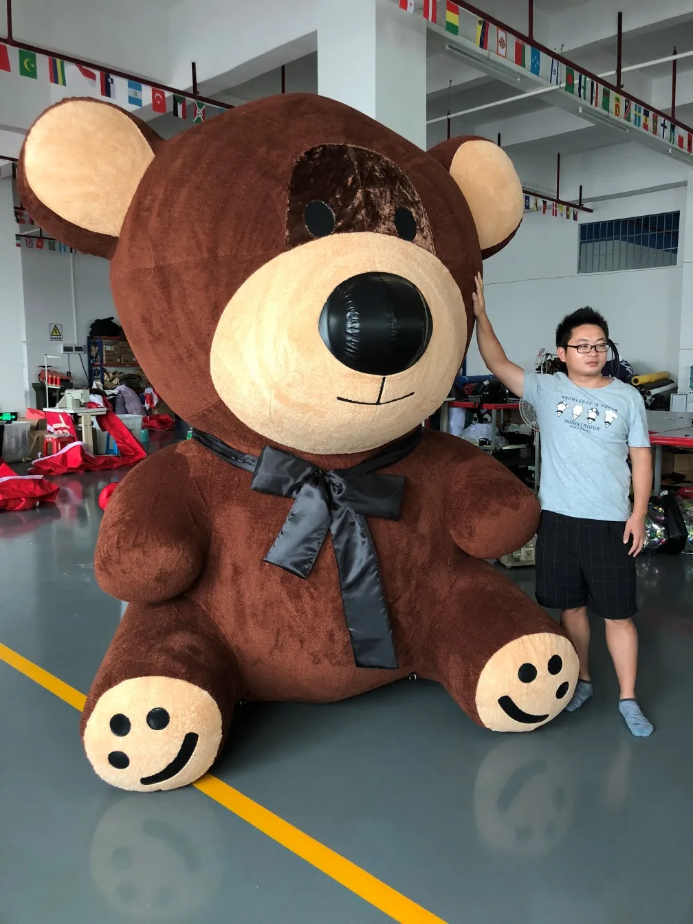 The giant brown plush teddy bear with a black satin bow, roughly two and a half times the height of the person standing beside it on the factory floor