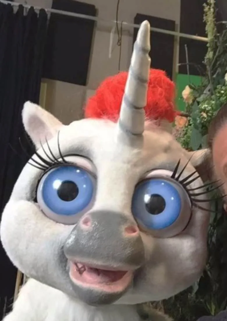 Close-up of the white unicorn character from the commercial: enormous blue eyes with long lashes, a grey muzzle, a red mane and a ribbed horn