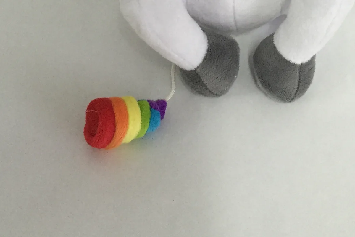 Close-up of the rainbow soft-serve: stacked plush rings in red, orange, yellow, green, blue and purple, on a white cord beside two grey hooves